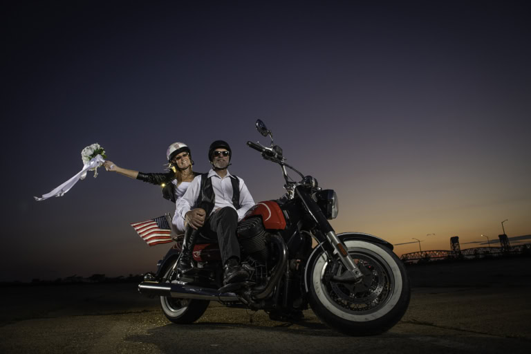 Two riders pose on a black motorcycle as an American flag waves beside them at sunset near a waterfront.