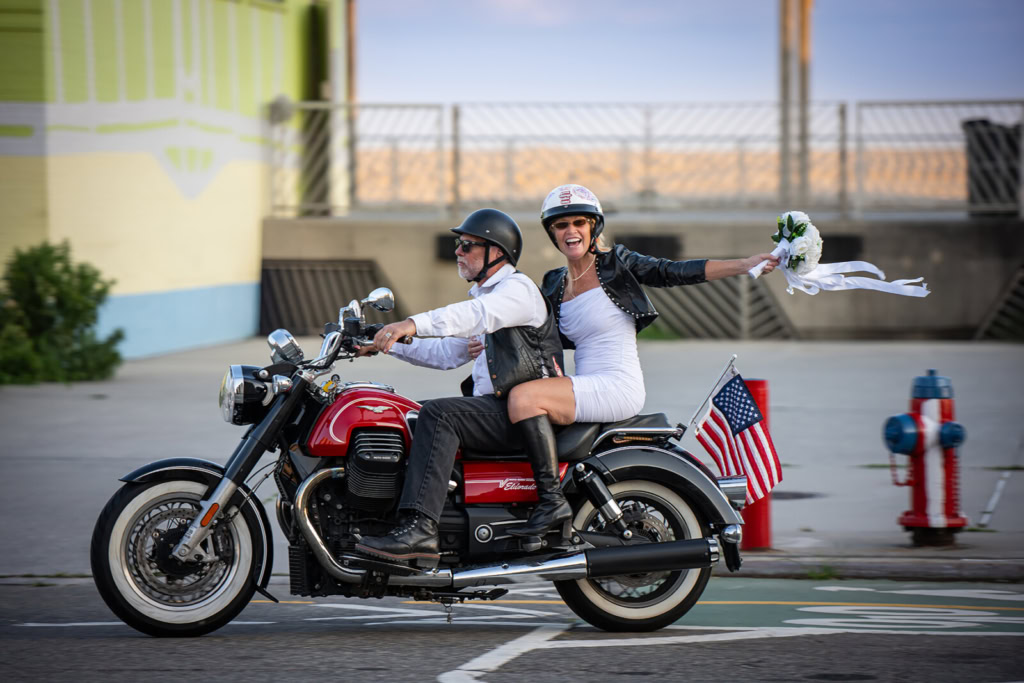 A bride in white and groom ride a red motorcycle past American flags on a city street during a wedding procession.